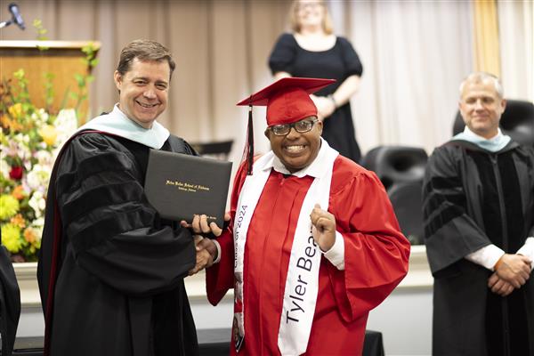 Dennis Gilliam, Ed.D. holds Tyler's diploma while shaking his hand. Both smile to camera.